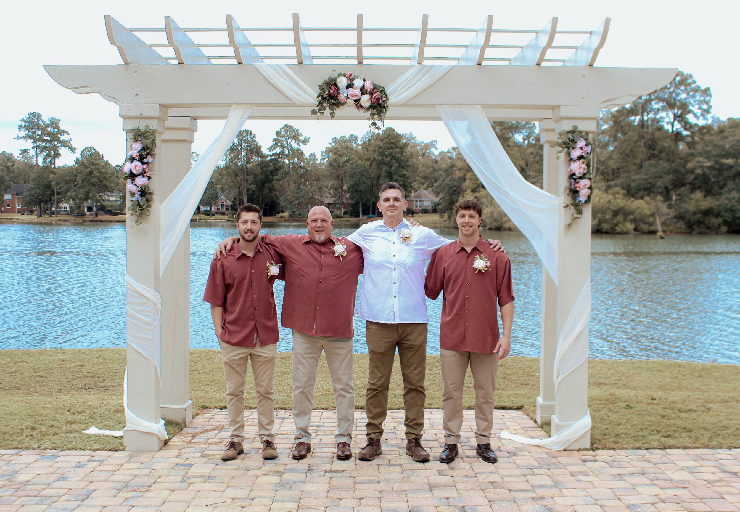 Four people in burgundy and white attire stand under a decorated wedding arch by a lake.