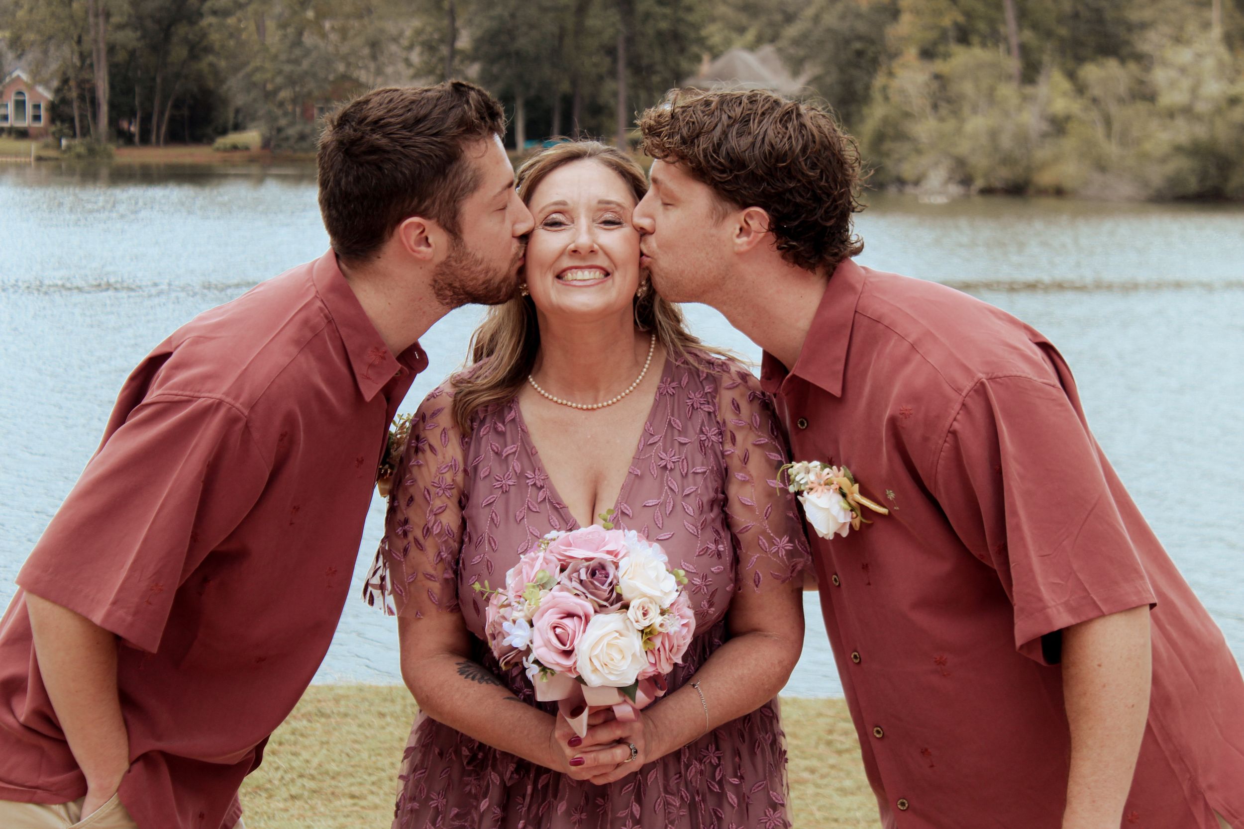 Two people kiss the cheeks of someone holding a pink and white bouquet under a wedding arch.