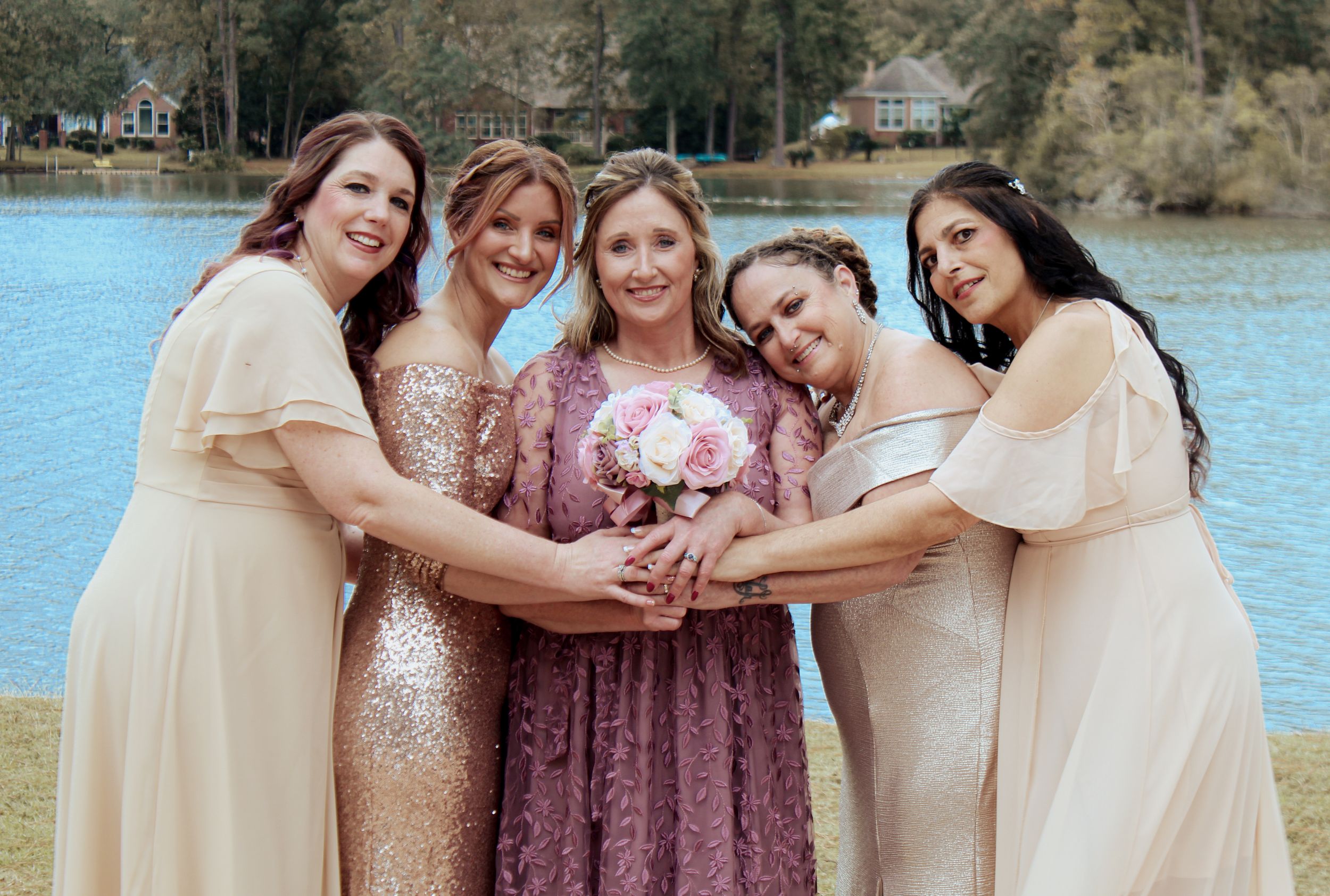 A group in cream and mauve dresses pose together holding a bouquet by a lake.