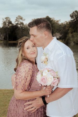 A couple embraces while holding a pink bouquet during their lakeside wedding photos.