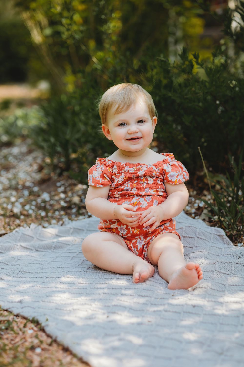 Baby in floral romper sitting on blanket in garden setting.