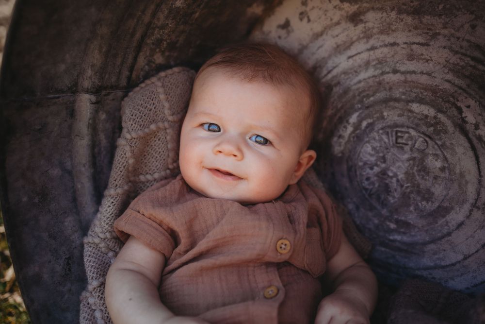 Baby wearing brown outfit smiling while sitting in rustic chair.