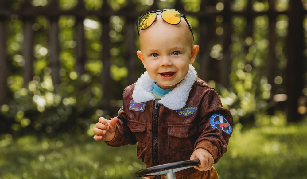 Baby in brown vest and sunglasses poses outdoors in dappled sunlight.
