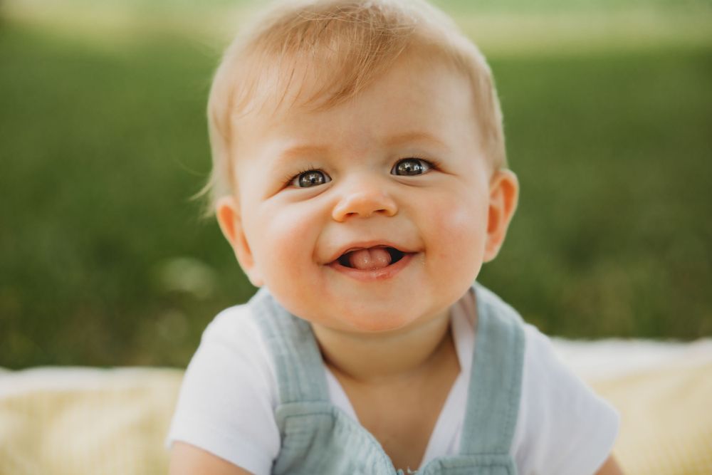 Baby in a white and gray outfit sits on grass with a bright cheerful expression.