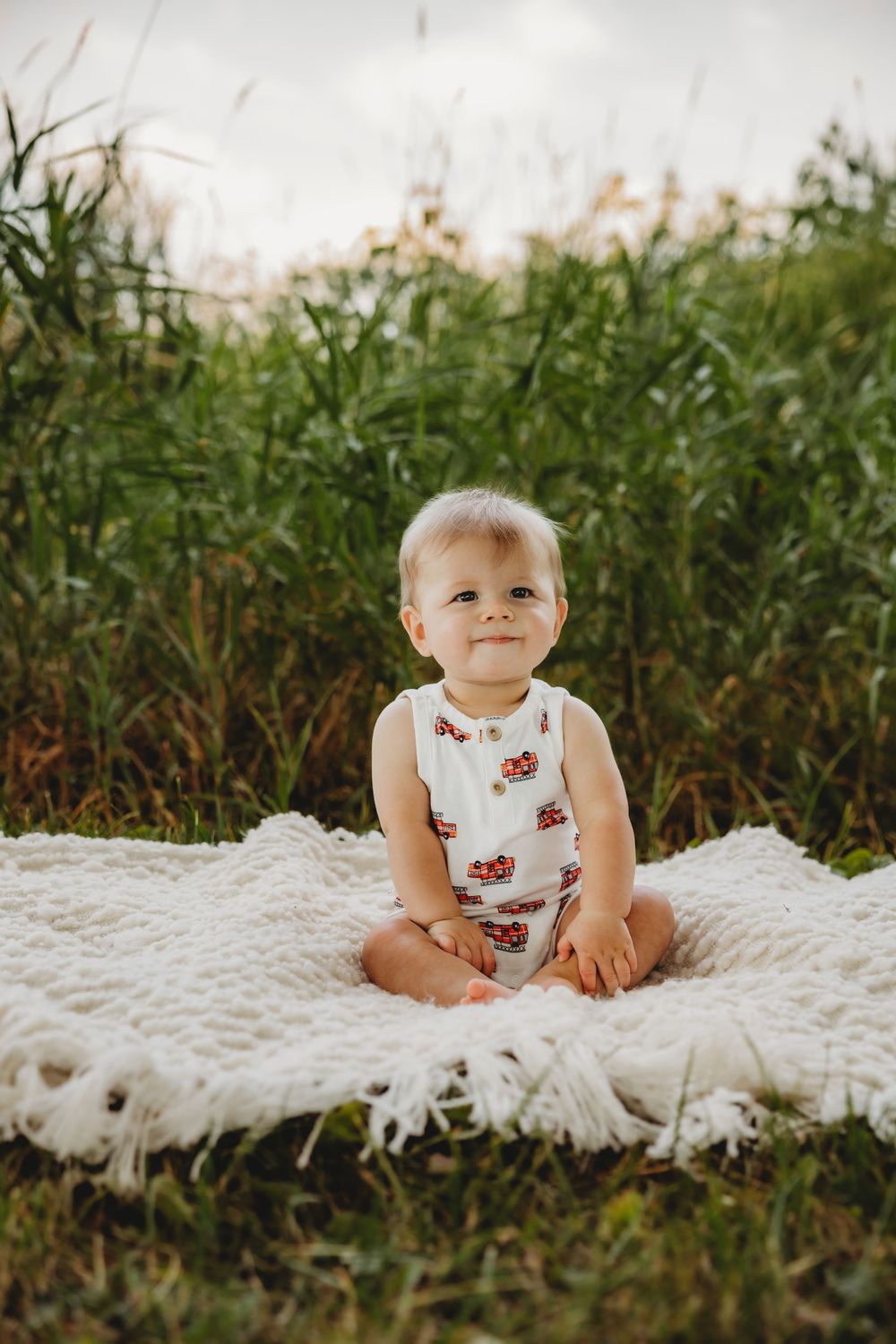 Baby wearing a white outfit with red details sits on a fluffy white blanket outdoors.