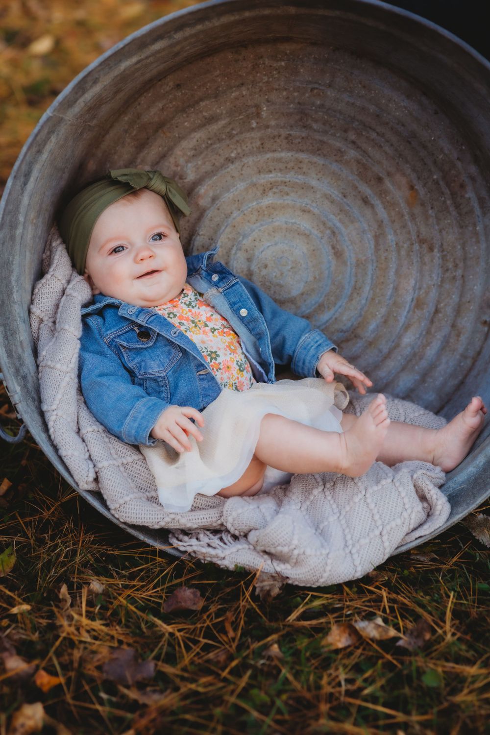 Baby wearing denim jacket resting in rustic metal tub with blanket.