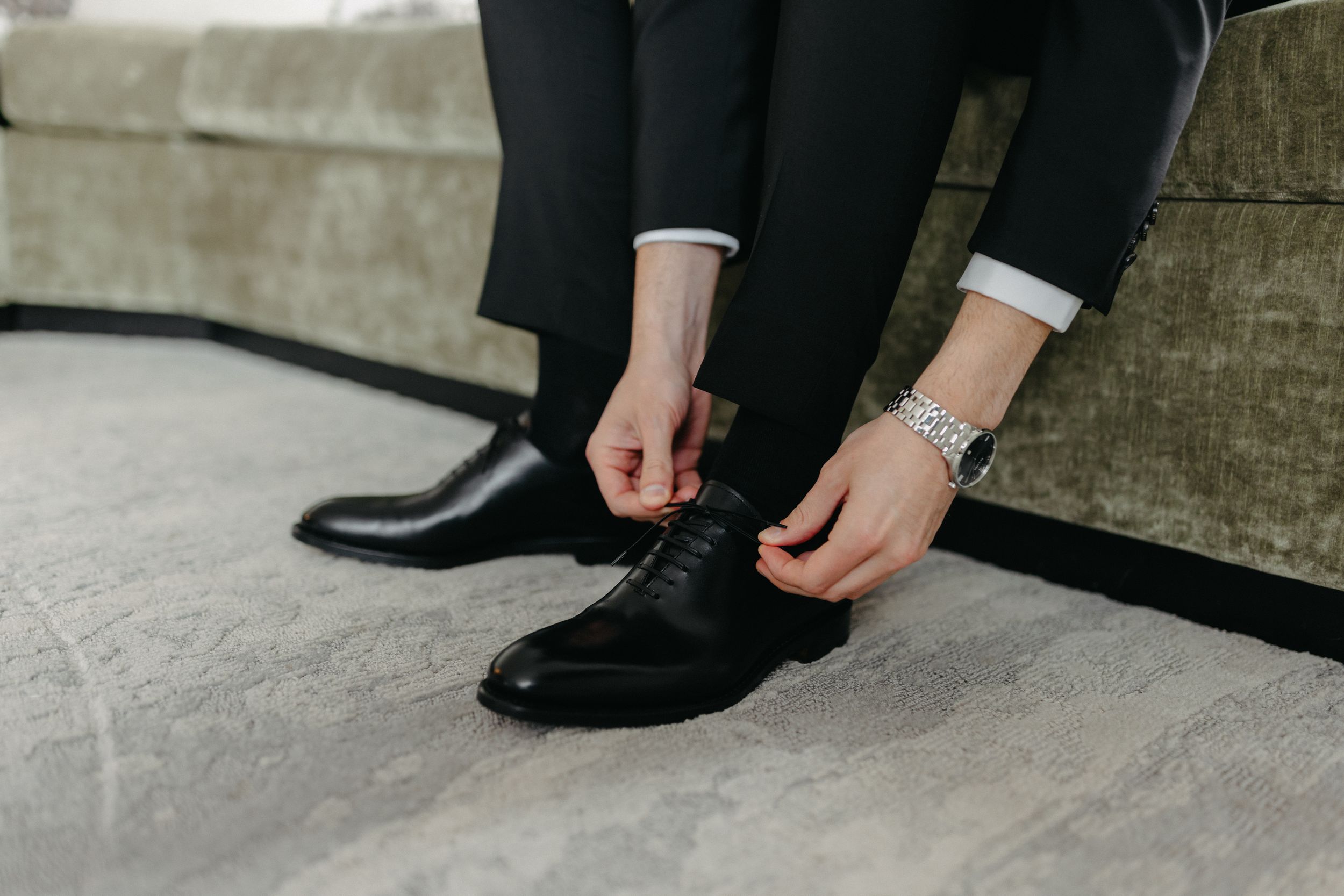 Close-up of hands adjusting black formal dress shoes on grey carpet.