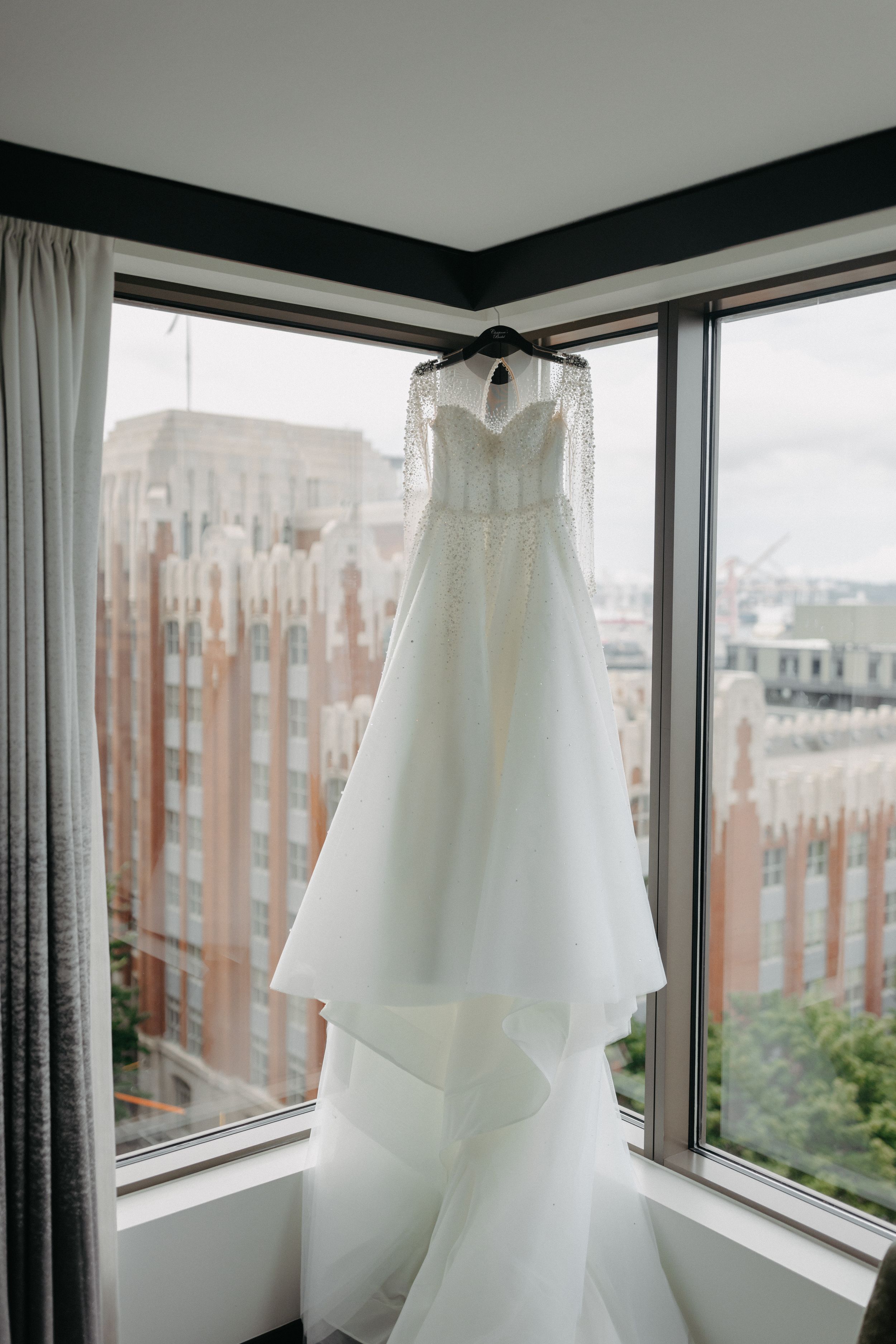 Elegant white wedding dress hanging in corner window with city building view in background.
