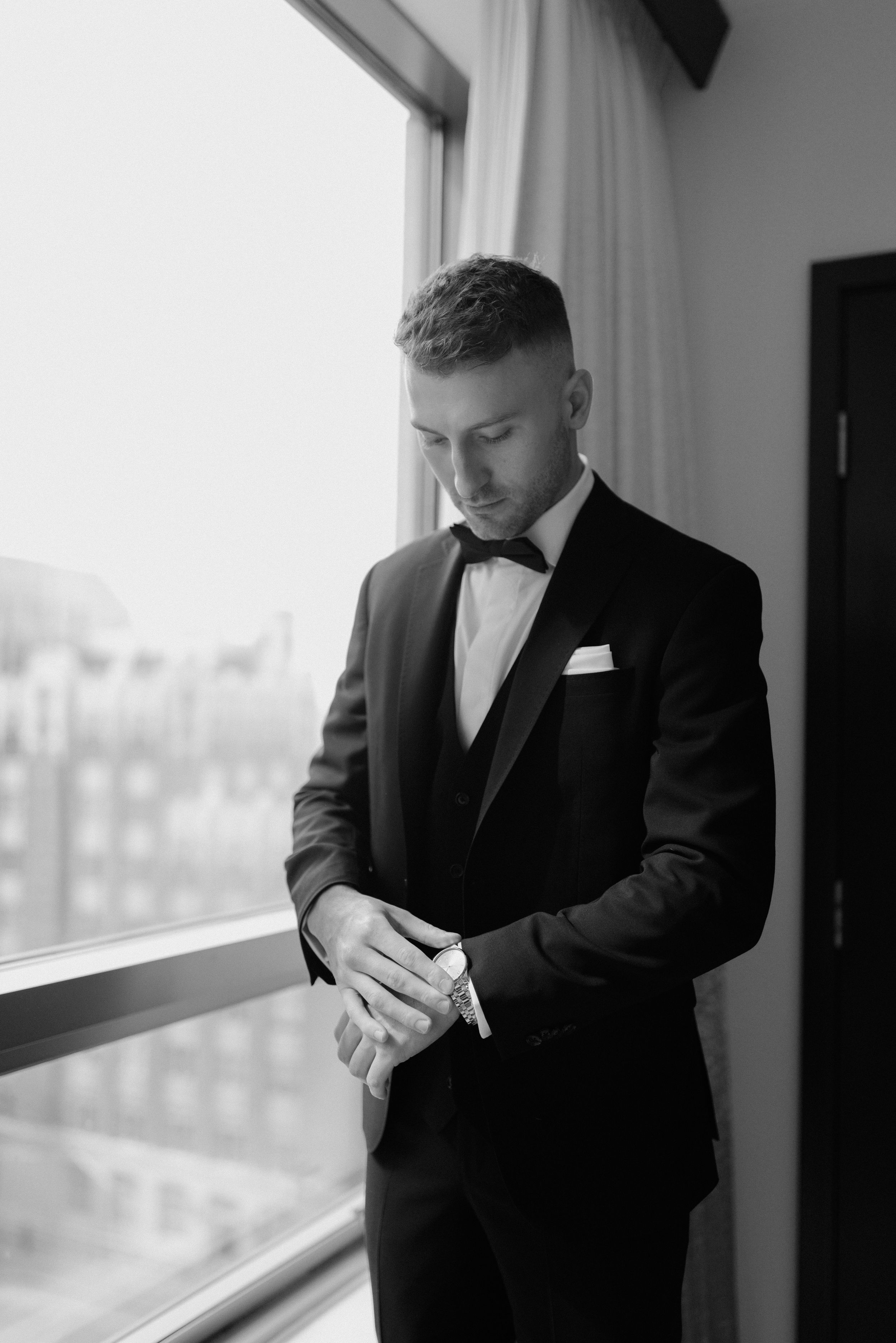 Figure wearing classic black tuxedo and bowtie looking at watch near window.