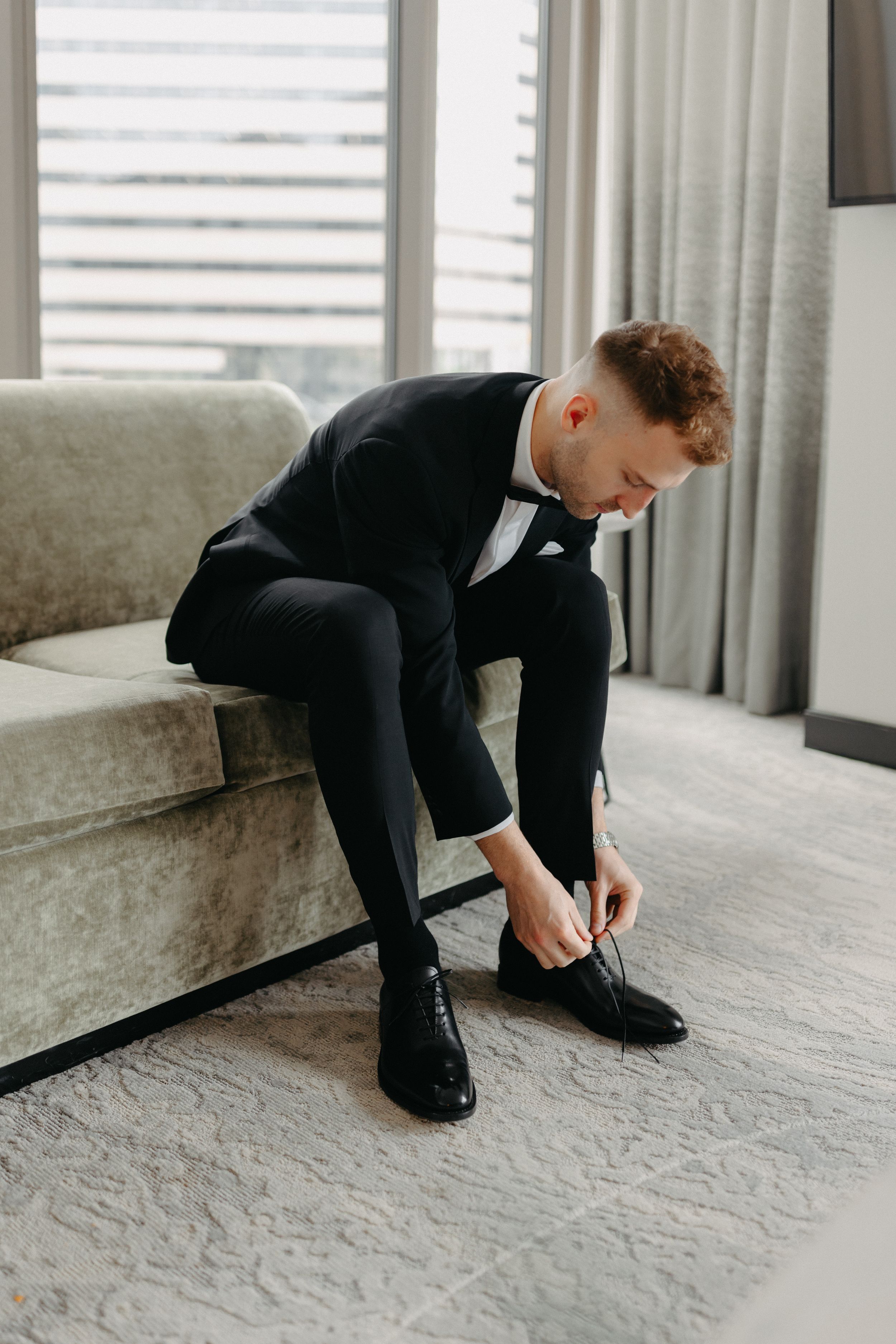 Someone in formal black tuxedo sitting on couch while tying dress shoes.