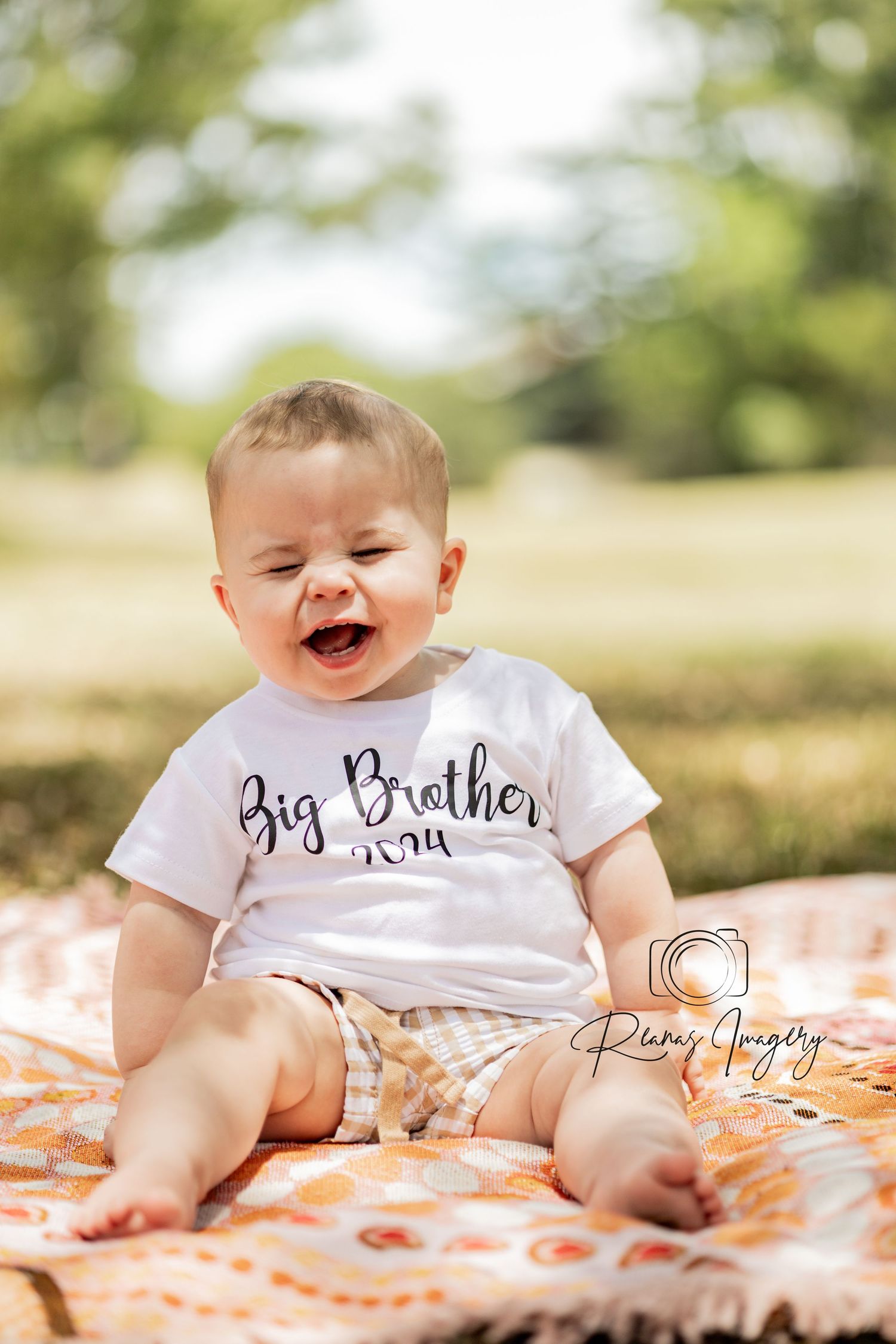 Happy baby boy sitting on a blanket outdoors wearing a Big Brother 2024 white shirt.