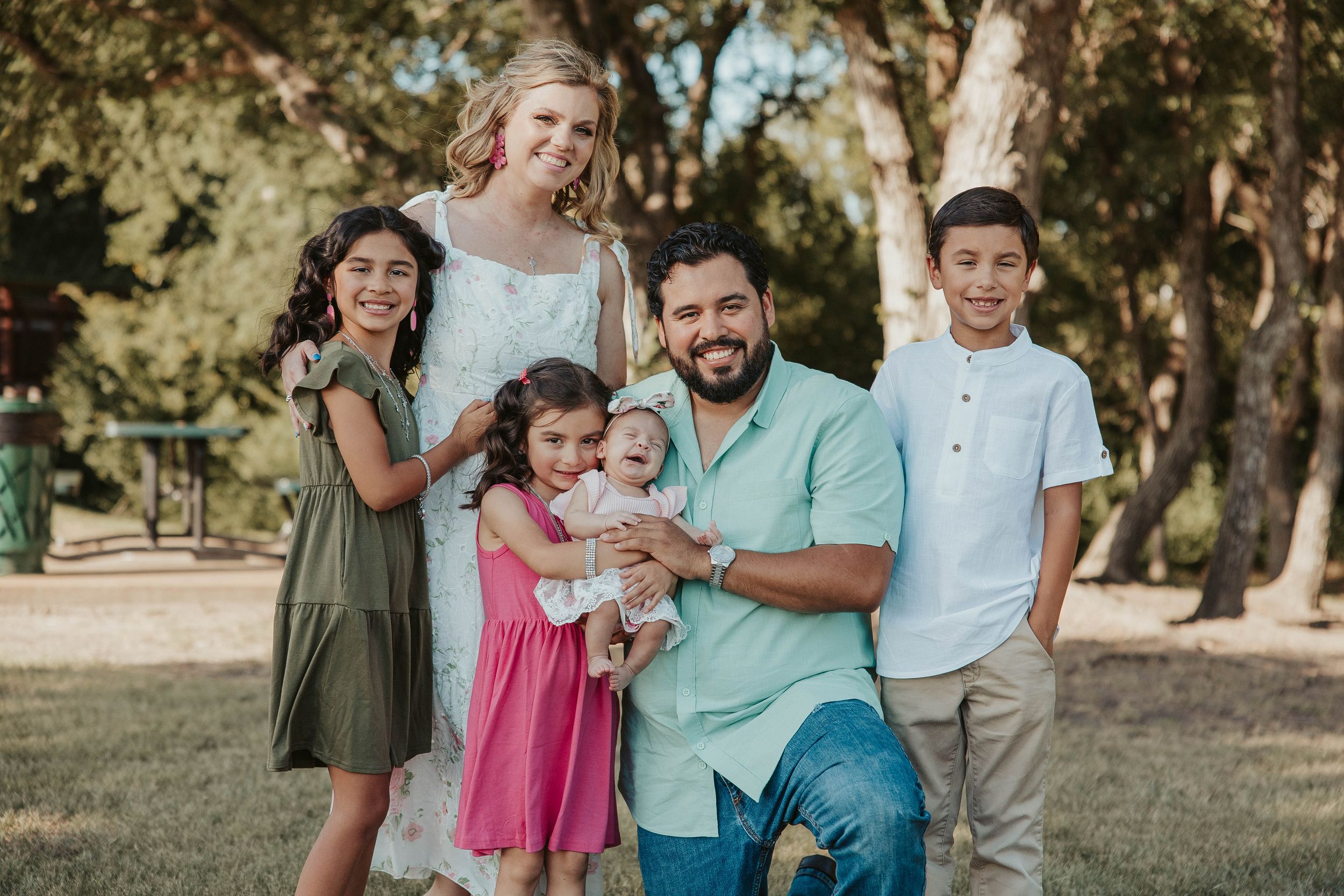 A happy family poses together outdoors for a portrait in casual attire against a tree-lined backdrop.