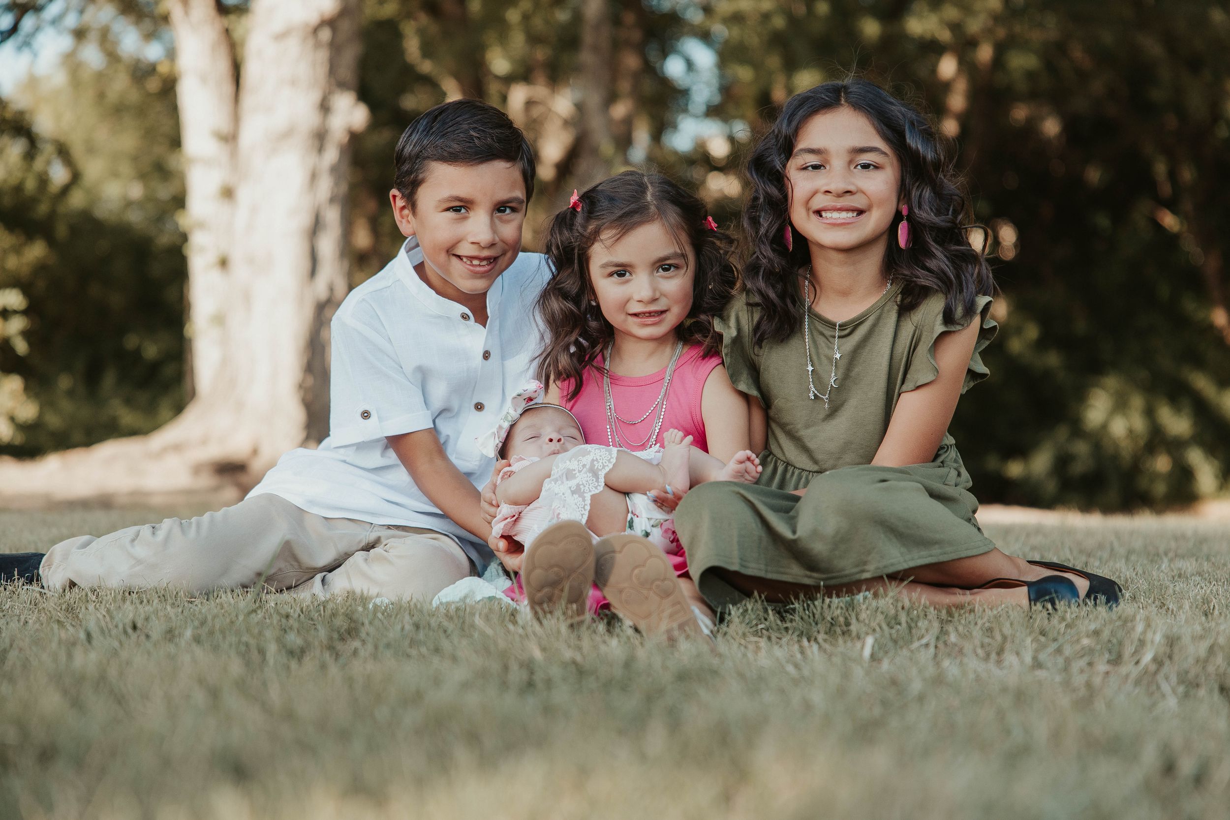 Three children sit together smiling on grass during a sunny outdoor photoshoot.