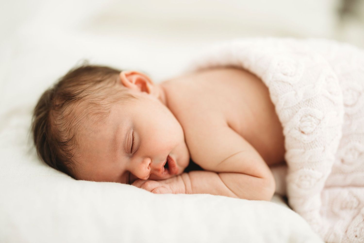 Peaceful sleeping newborn baby wrapped in soft white blanket resting on white bedding.