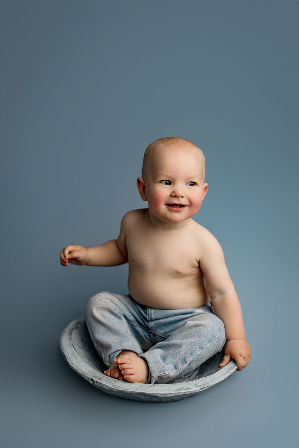 Baby in gray pants sits happily on round cushion against blue studio background.