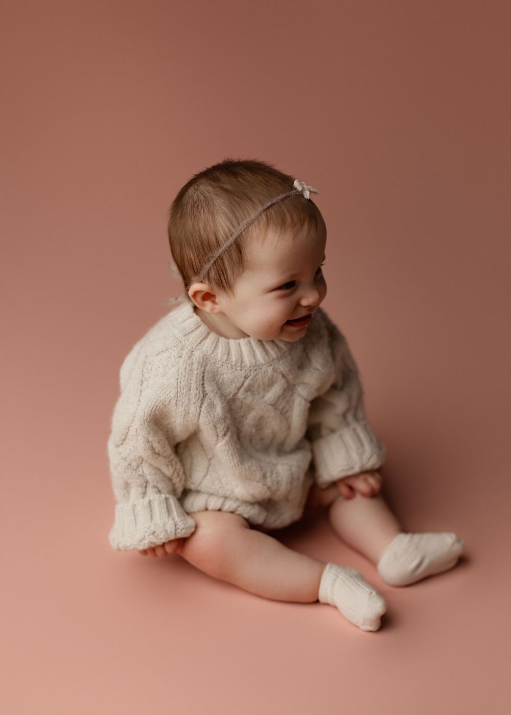 Baby in cozy cream sweater smiles while sitting on pink backdrop.