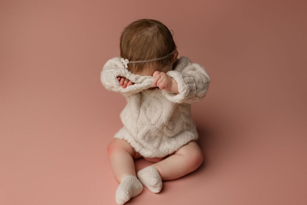 Baby in white ruffled outfit with bow sits against pink background.
