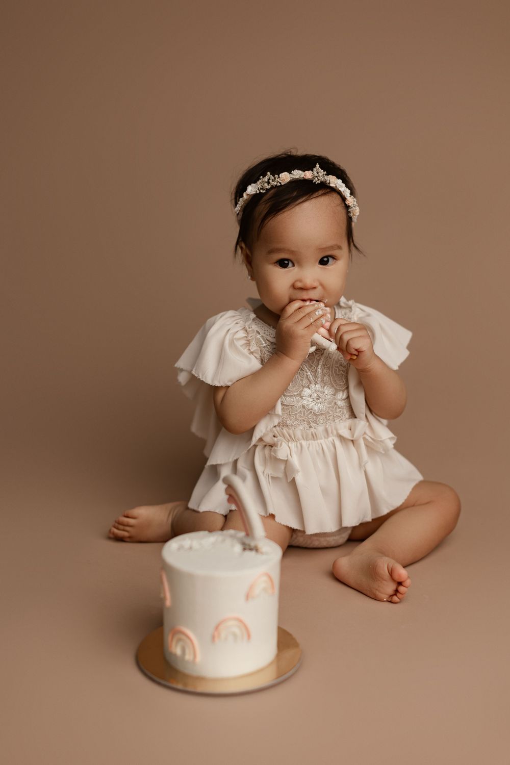 Baby in white lace dress reaching for decorative cake on brown background.