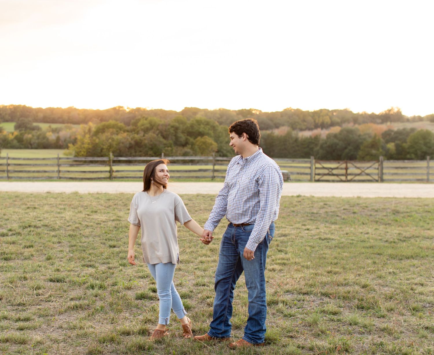 Couple holding hands while walking in a rural field with wooden fences at sunset.