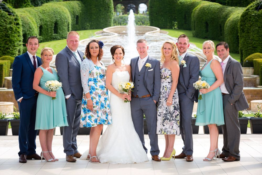 Wedding party in blue and mint green attire posed in front of manicured garden hedges.