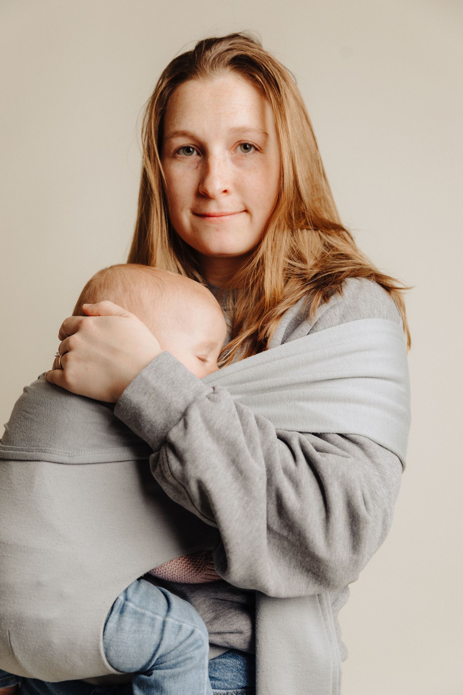 Parent in grey sweatshirt holding sleeping baby against neutral background.