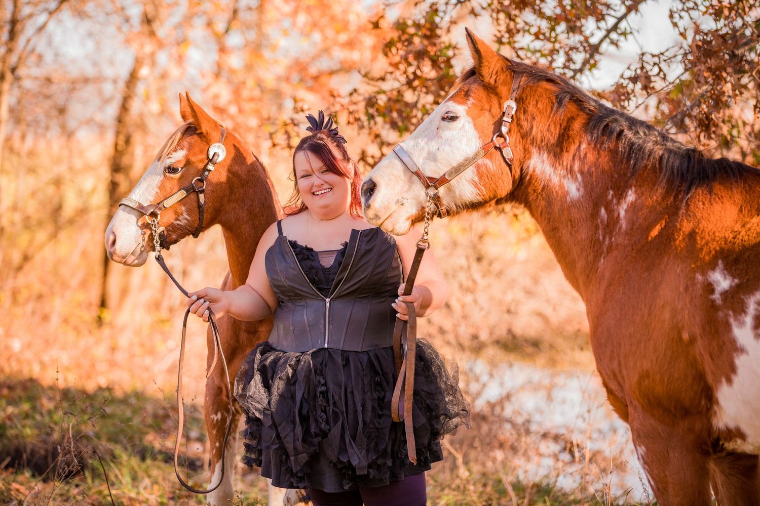 Person in black dress poses between two majestic brown horses during golden hour sunset.
