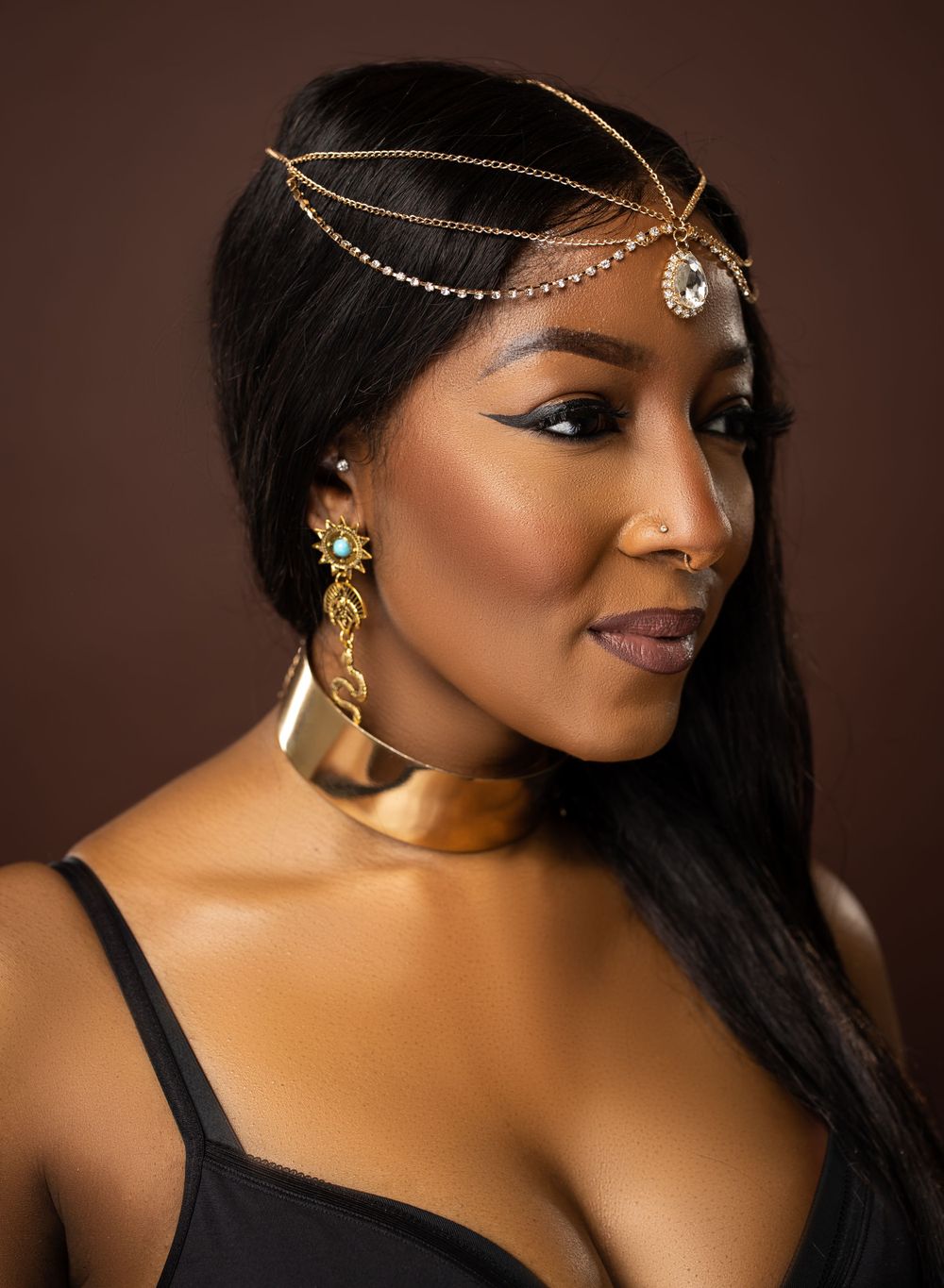 Elegant portrait featuring ornate gold headpiece, choker and dangling earrings against brown background.