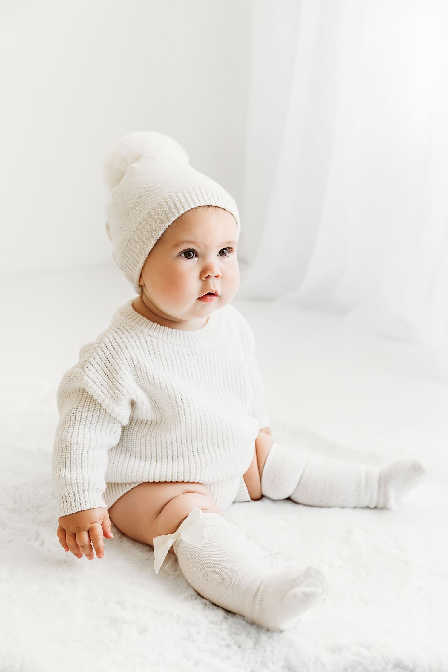 Baby in a white winter hat with pom pom, sweater onesie, and knee high socks