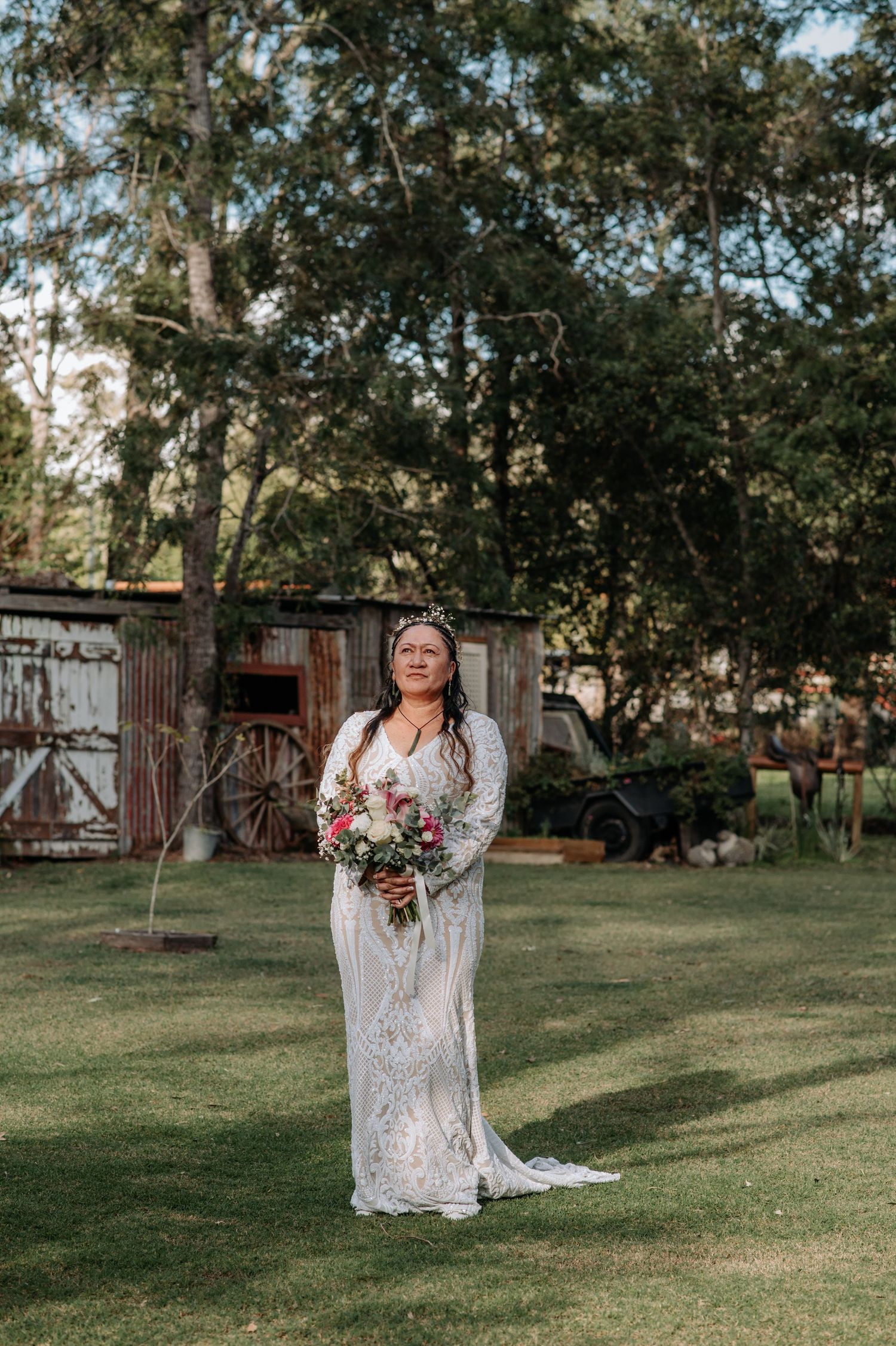 Bride in lace gown poses with bouquet in rustic outdoor setting.
