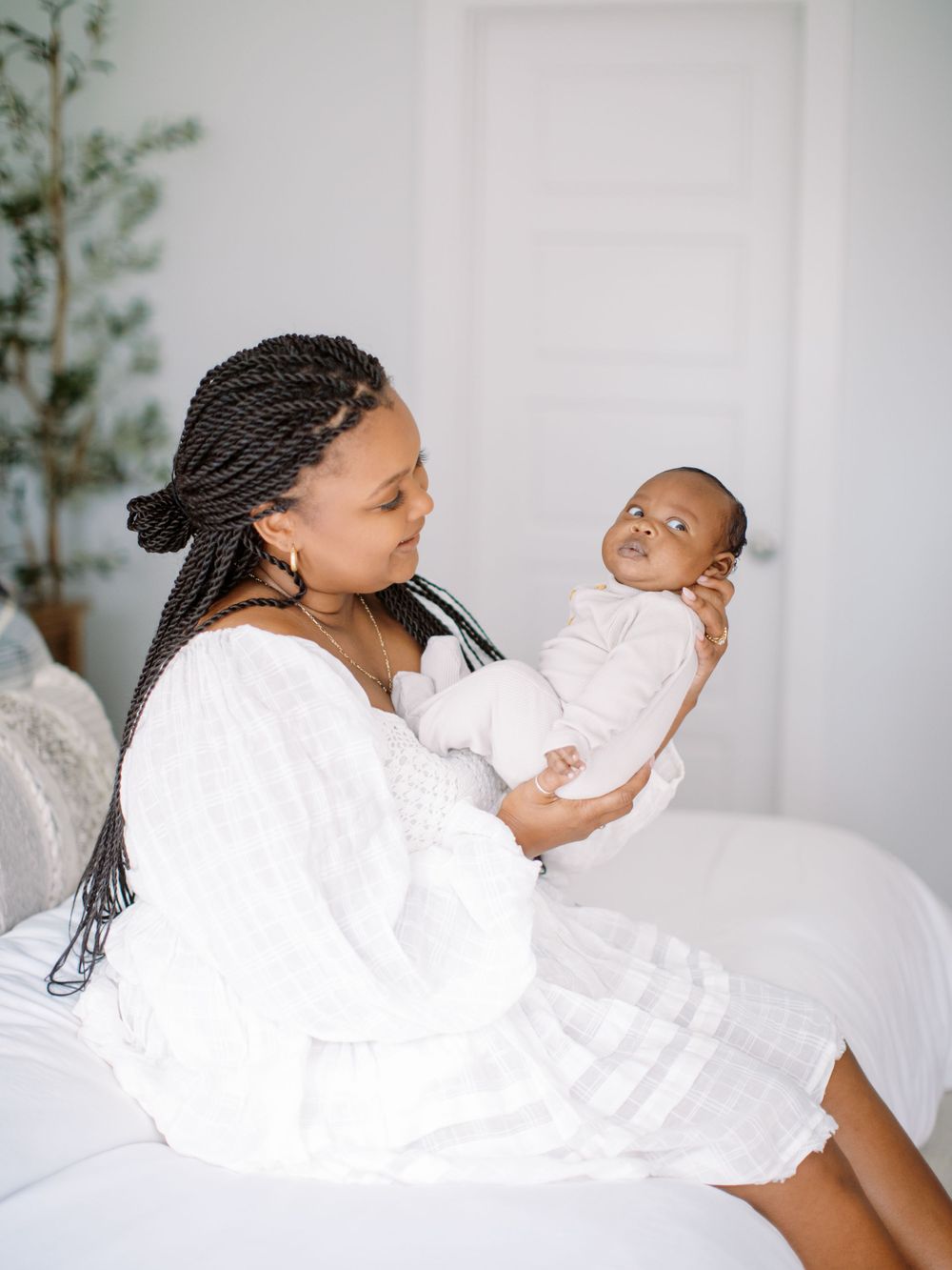 A tender moment between parent and infant wearing white captured in a bright, minimalist bedroom setting.
