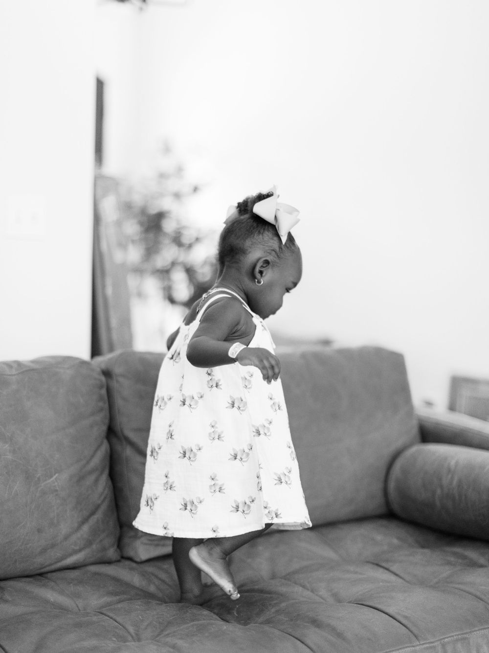 Black and white photo of toddler in floral dress on couch.