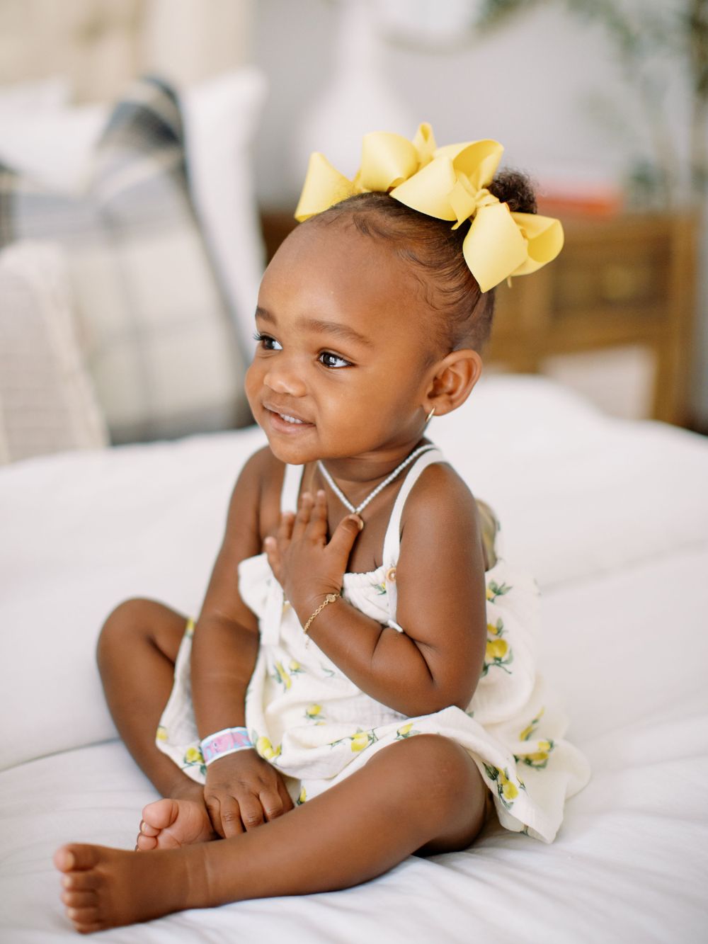 Young child in floral dress with yellow bow sitting on white couch.