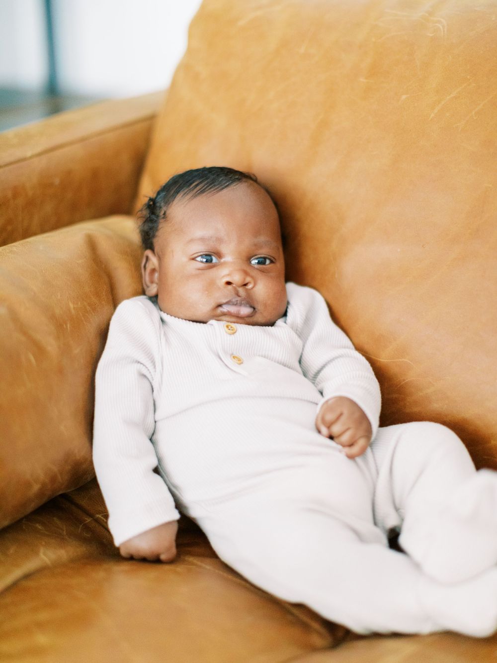 Baby in white onesie reclines on tan leather sofa.