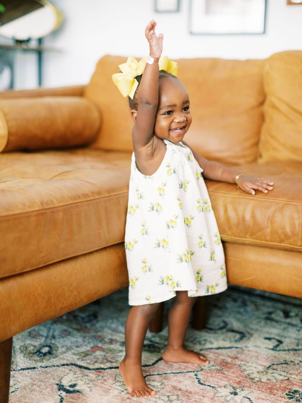 Happy toddler in floral dress dances by tan leather couch.