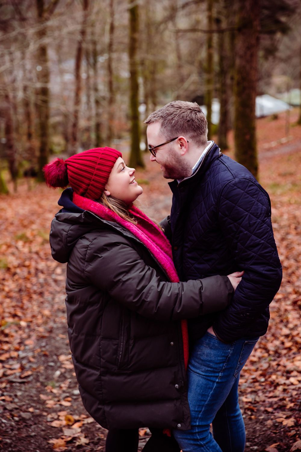 A romantic winter couple embraces on fallen leaves in the forest wearing warm winter clothing.