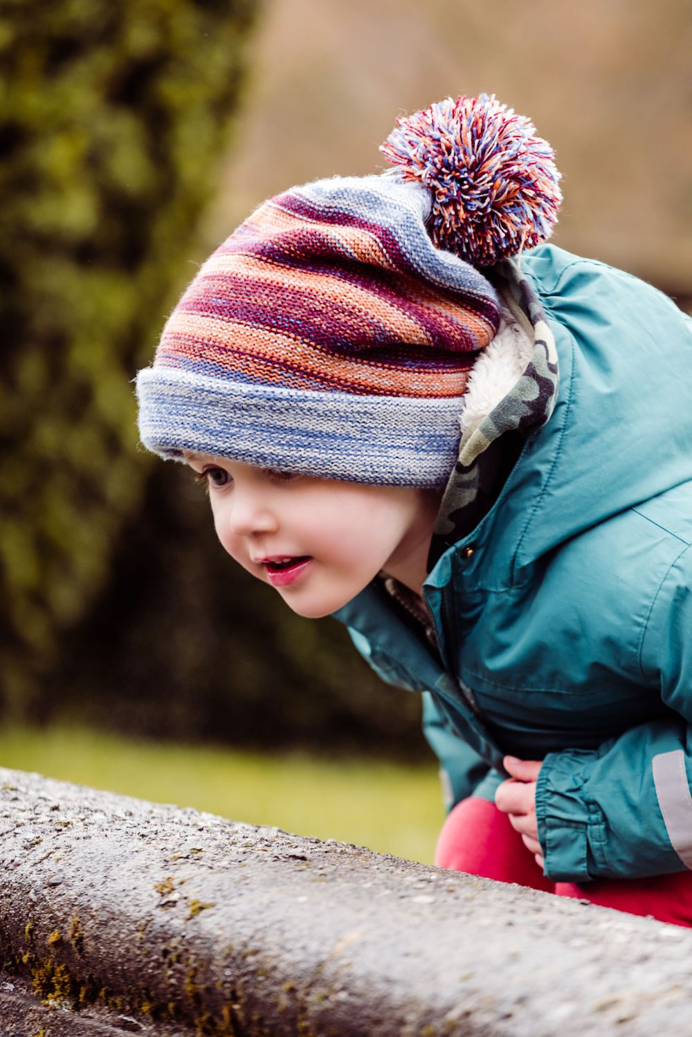 Young explorer in winter attire leans forward to examine something while wearing a colorful striped beanie with pom-pom.