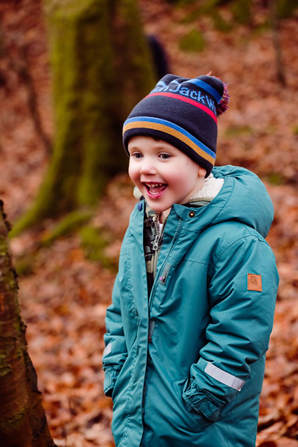 Happy child in a teal winter coat and patterned beanie smiles joyfully in the forest.