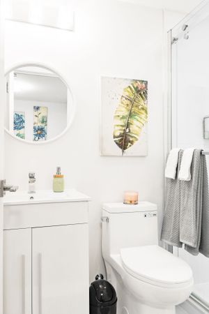 Clean white bathroom with round mirror, leaf artwork, and gray towels.