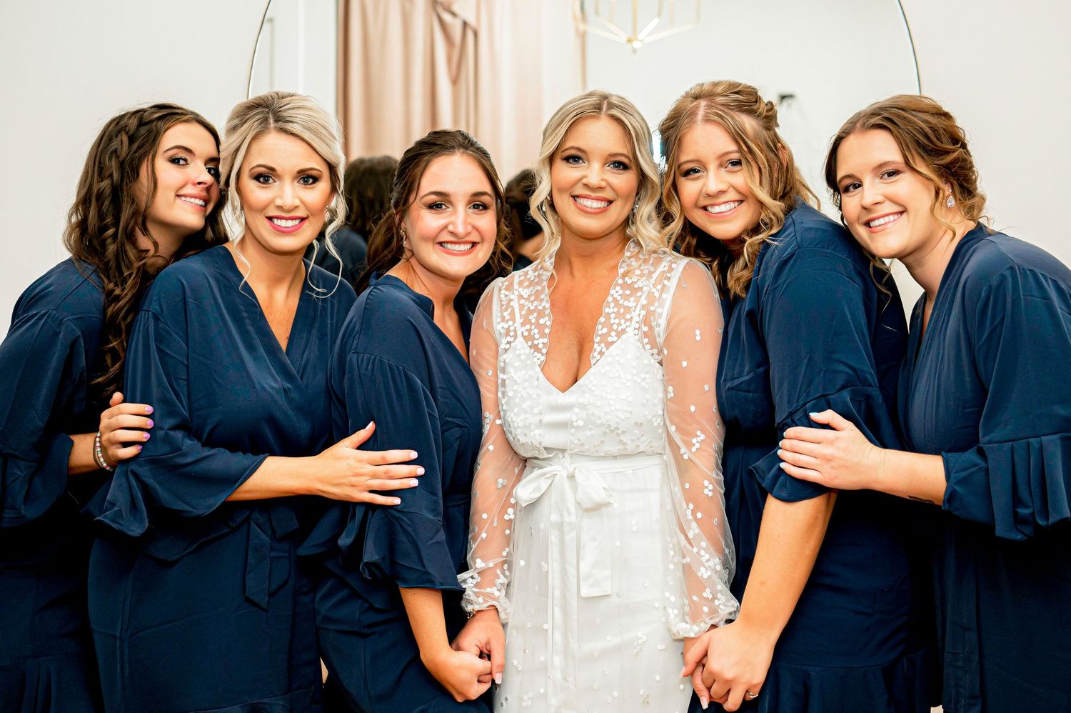 Bridesmaids in navy robes gather around bride in white for celebratory photo.