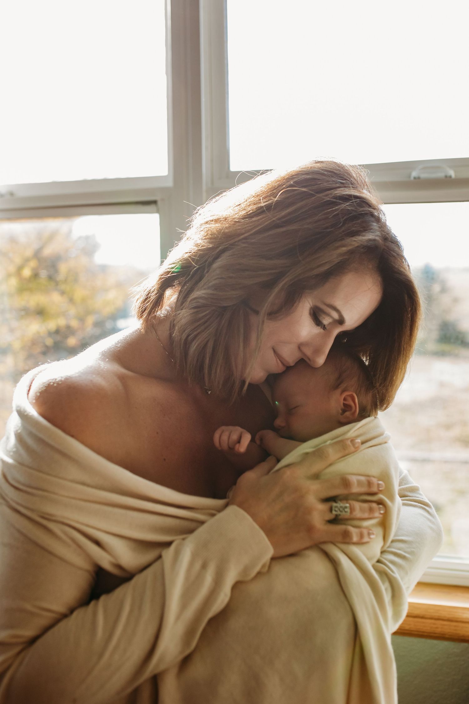 Tender moment by window with baby wrapped in beige blanket.