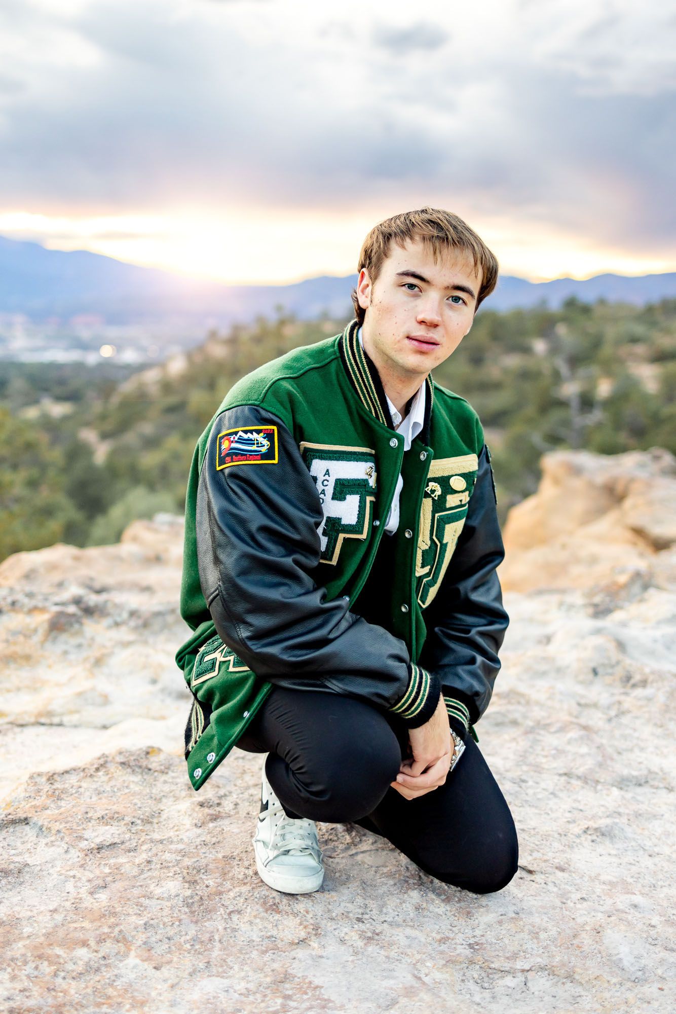 Senior guy in letter jacket kneels on the rocks at sunset at his senior session at Palmer Park in Colorado Springs.