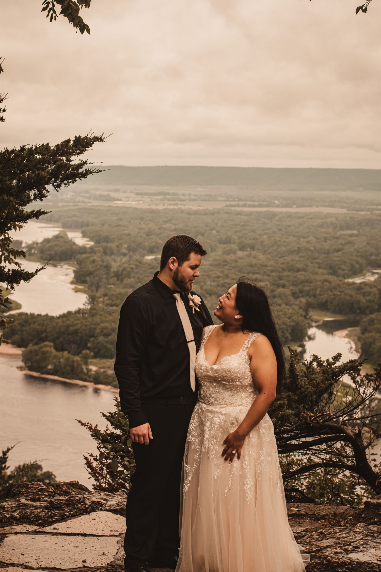 Couple in wedding attire sharing intimate moment on clifftop overlooking river valley.
