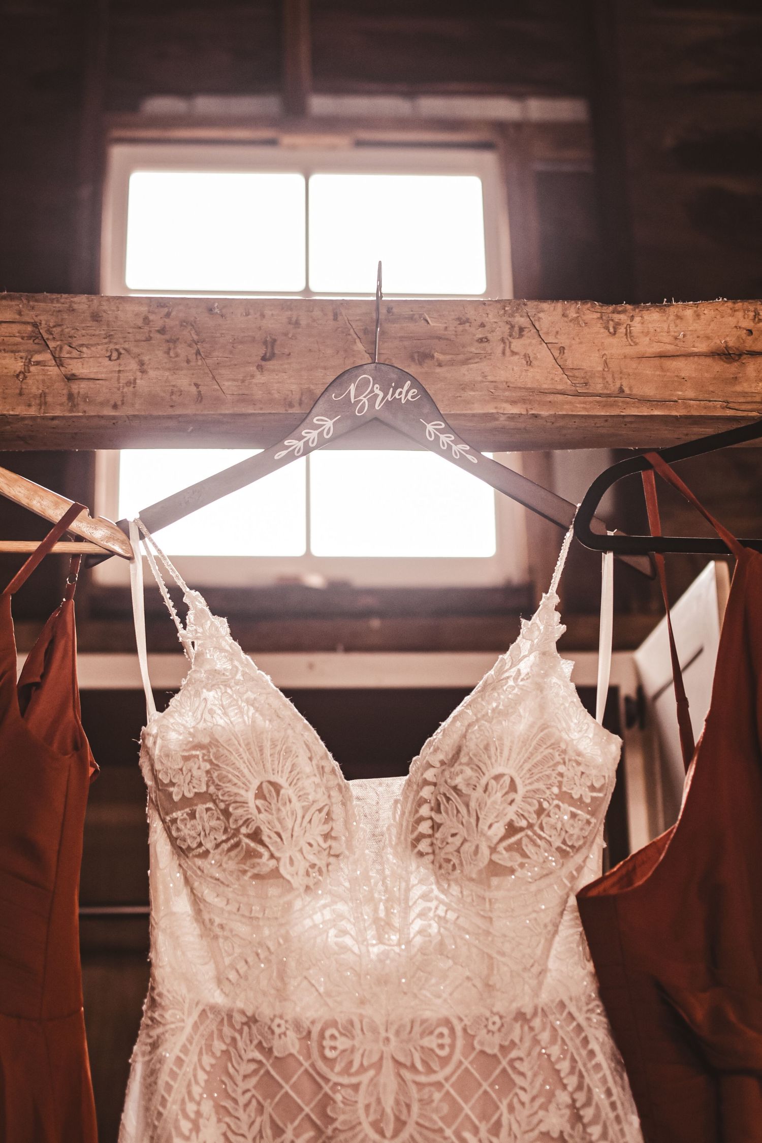 Delicate lace wedding dress hangs on wooden beam in rustic setting with natural window light.