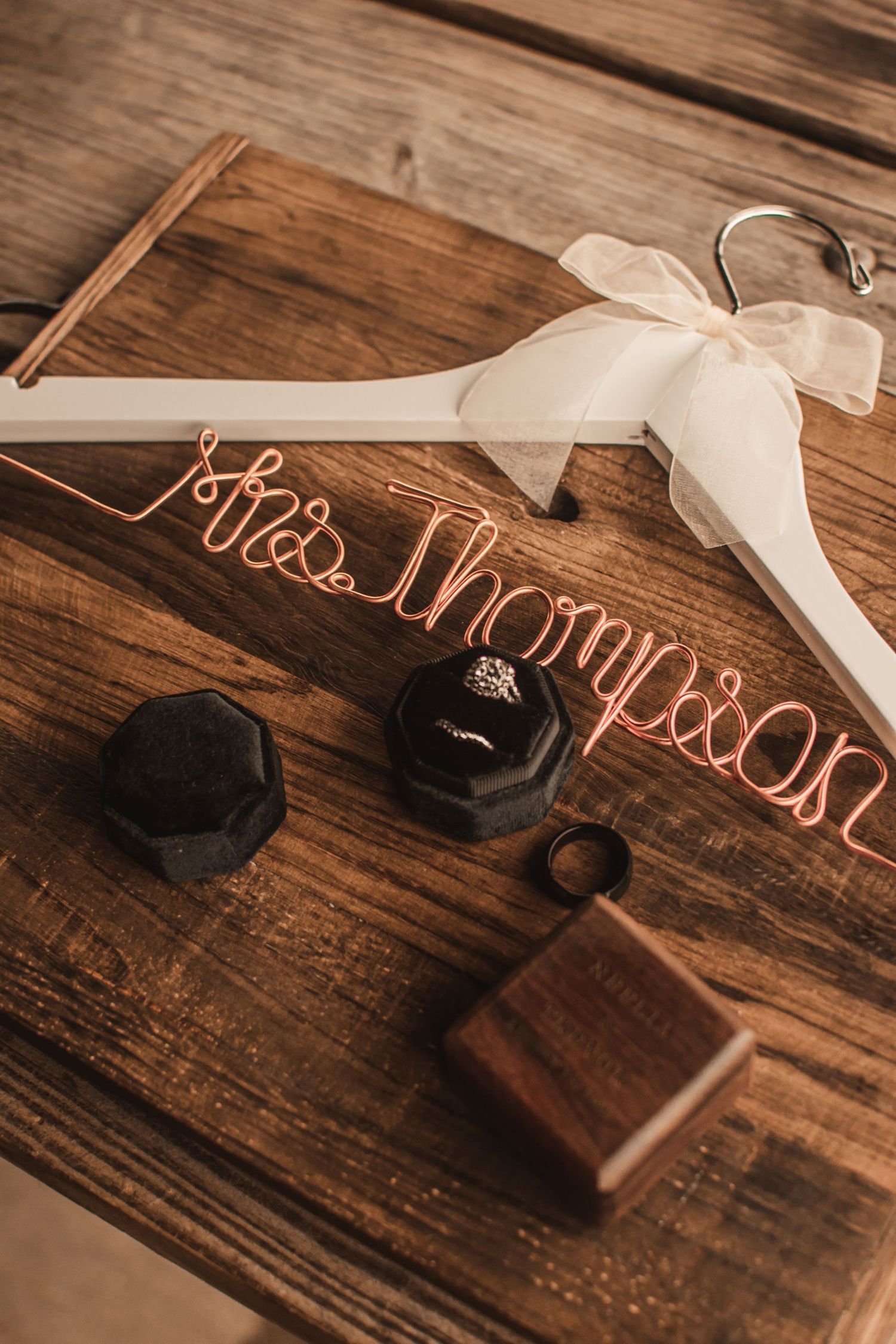 Wedding details with personalized wire hanger, white ribbon, and rings on wooden surface.