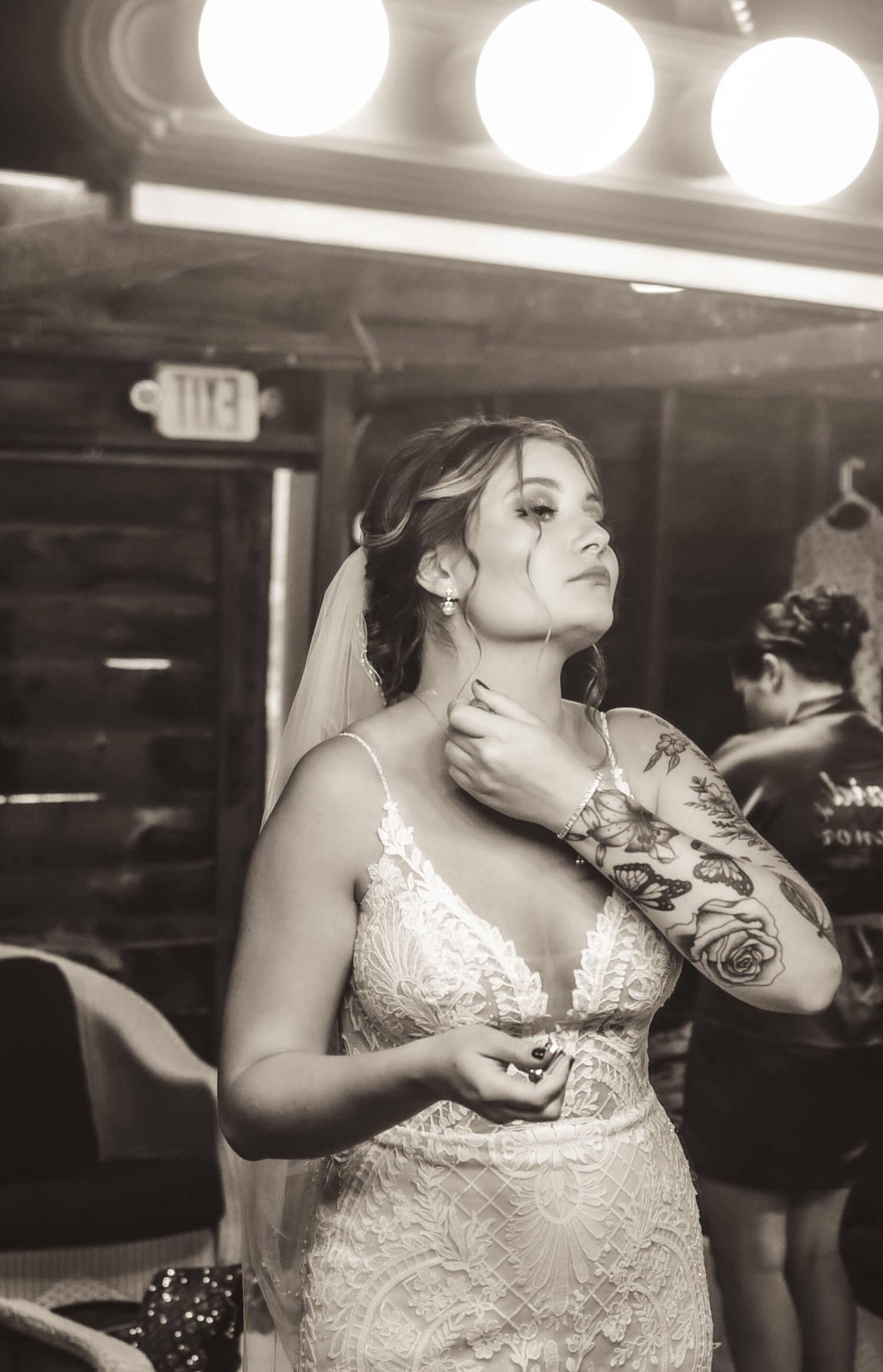 Black and white portrait of bride in lace dress with visible tattoo under vanity lights.
