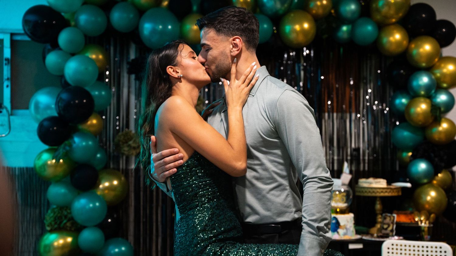 Couple sharing romantic moment at party with teal and gold balloon arch backdrop.