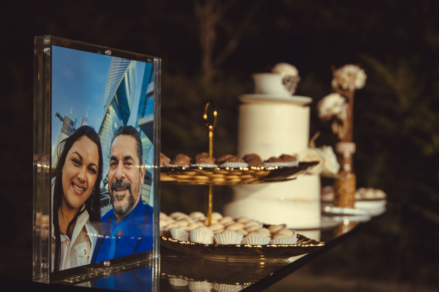 Wedding reception dessert table with white cake and framed photos in dim lighting.