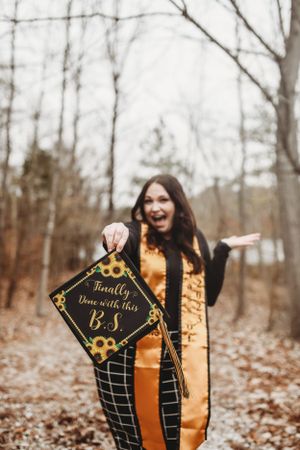 Graduate holding decorated mortarboard with 'B.S.' and sunflower design in wooded setting.