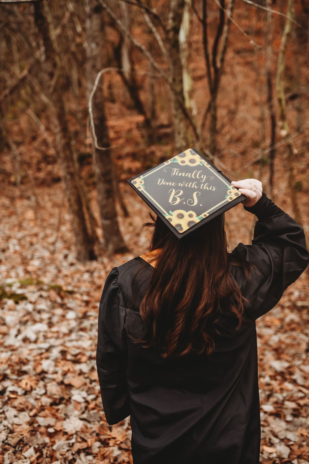 Graduation cap decorated with sunflowers photographed from behind in autumn forest setting.