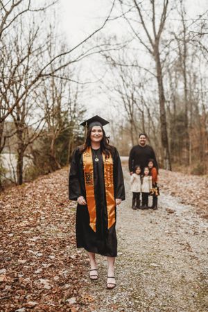 Graduate in black cap and gown with orange stole walking on leaf-covered path.
